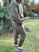 Load image into Gallery viewer, Person wearing an olive green tracksuit standing outdoors on a grassy area with trees in the background.