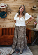 Load image into Gallery viewer, A woman modeling a bohemian-style outfit consisting of a white short-sleeve tie-front blouse and a brown maxi skirt with a white chain-link pattern, standing in front of a rustic wood-paneled wall with a brown velvet sofa.