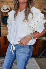 Load image into Gallery viewer, A woman wearing a white ruffled short-sleeve top with a front zipper and drawstring hem, paired with blue jeans, in a rustic indoor setting with wood paneling and an orange chair.