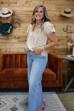 Load image into Gallery viewer, A woman modeling a light beige short-sleeve blouse with ruffles and light blue flare jeans, standing in front of a rustic wood-paneled wall with a brown velvet sofa and cowboy hats in the background.