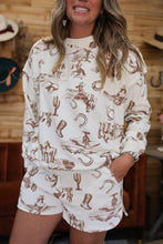 Load image into Gallery viewer, A woman wearing a matching cream-colored lounge set with a brown western-themed print featuring cacti, horseshoes, boots, and cowboys, set against a rustic wooden wall background.