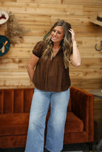 Load image into Gallery viewer, A woman modeling a brown short-sleeve button-up top with textured vertical pleats, paired with light-wash blue flare jeans, standing in front of a wooden wall and an orange-brown velvet sofa.