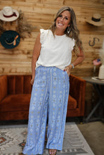 Load image into Gallery viewer, A woman modeling a white sleeveless top and light blue wide-leg pants with a white geometric pattern, standing in a rustic room with wood-paneled walls, a brown velvet chair, and hats hanging on the wall.