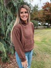 Load image into Gallery viewer, Woman wearing a brown hoodie and blue jeans standing outdoors with trees in the background