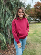 Load image into Gallery viewer, Woman wearing a maroon hoodie and blue jeans standing outdoors with trees and grass in the background