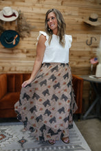 Load image into Gallery viewer, A woman modeling a white ruffle-sleeve top and a long beige skirt with a black horse pattern and small orange floral accents, standing in a rustic room with wood-paneled walls and cowboy hats.
