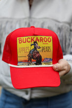 Load image into Gallery viewer, Red trucker hat with a vintage-style label design, held by a person wearing a light gray jacket.
