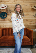 Load image into Gallery viewer, A woman modeling a white long-sleeve button-up shirt with a colorful cowboy and western-themed print, paired with blue jeans, standing in front of a rustic wood-paneled wall with an orange-brown velvet sofa.
