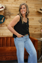 Load image into Gallery viewer, A woman modeling a black sleeveless button-front top and light-wash blue jeans in a rustic indoor setting with a wood-paneled wall and an orange-brown velvet sofa.

